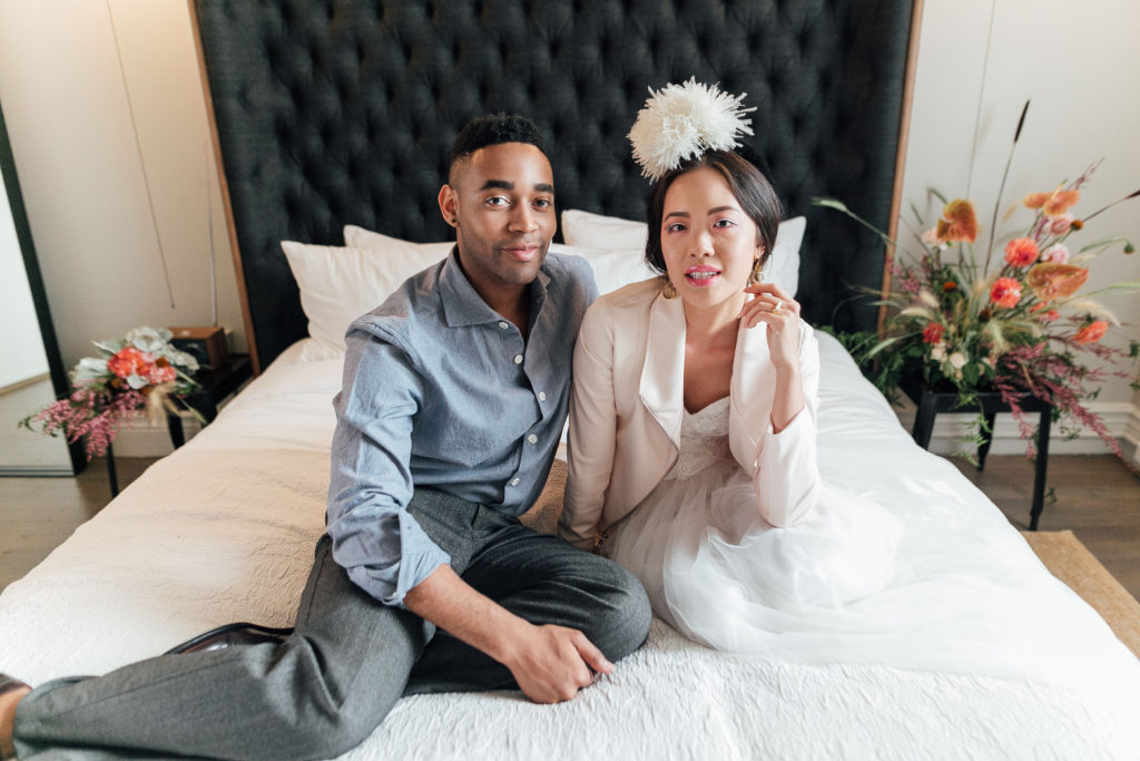 Desmond and Chanel sitting on bed at Hotel G wearing ethically-made Kali Made Garments menswear, Celia Grace wedding dress, Brevity Brand jacket, Soko jewelry, and vintage accessories