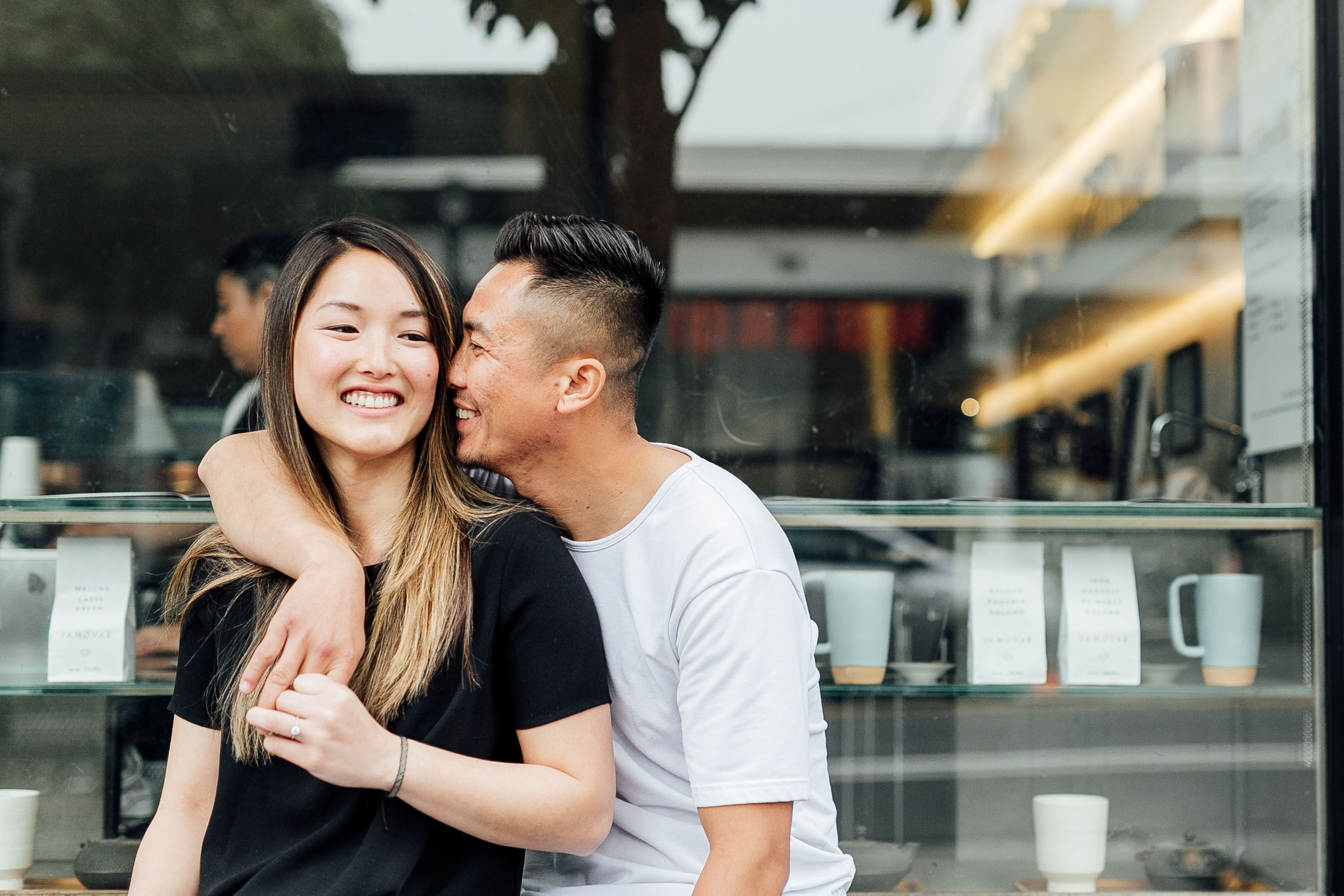 couple stands in front of a storefront window, fiancée smiles