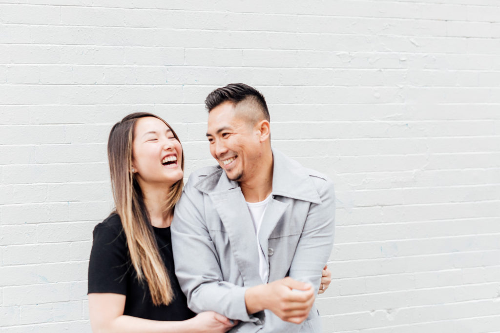 engaged couple laughs together in front of white wall 