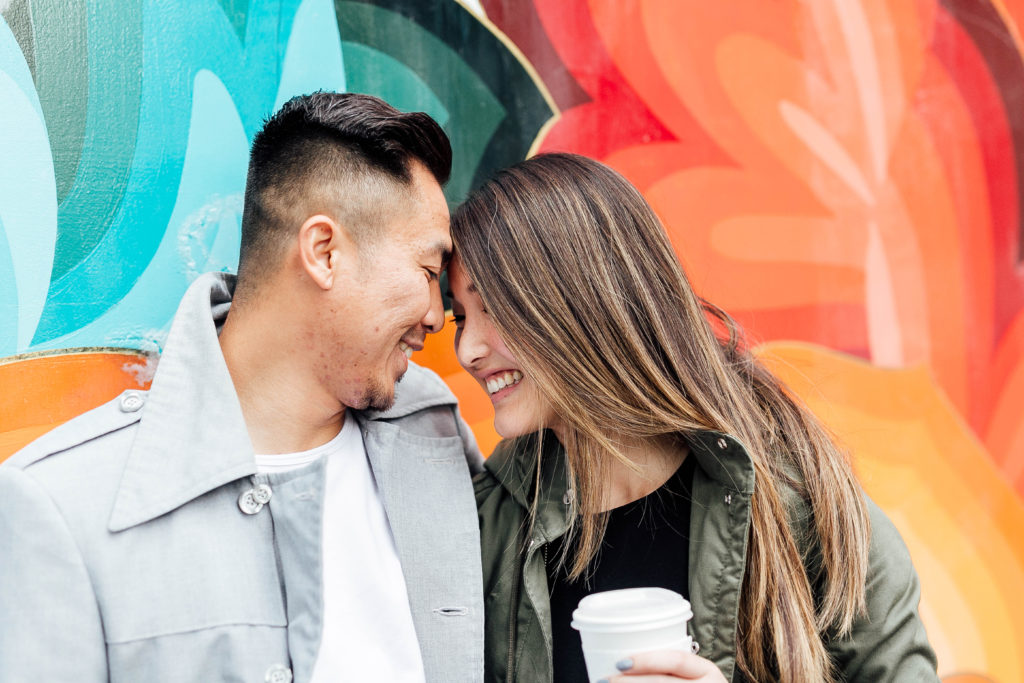 engaged couple lean lovingly on each other in front of colorful wall