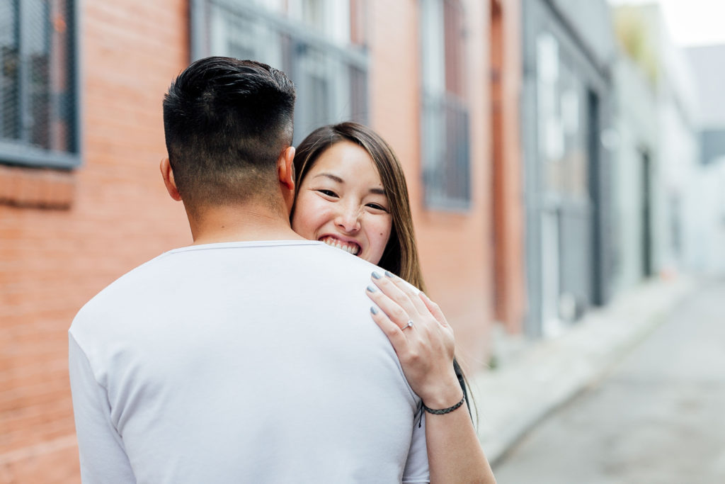 couple embraces in alley, fiancée smiles while hugging her partner