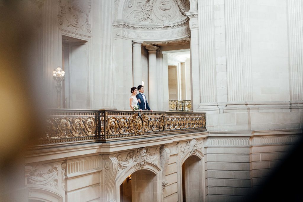 Bride and groom stand on elegant balcony, looking out at City Hall