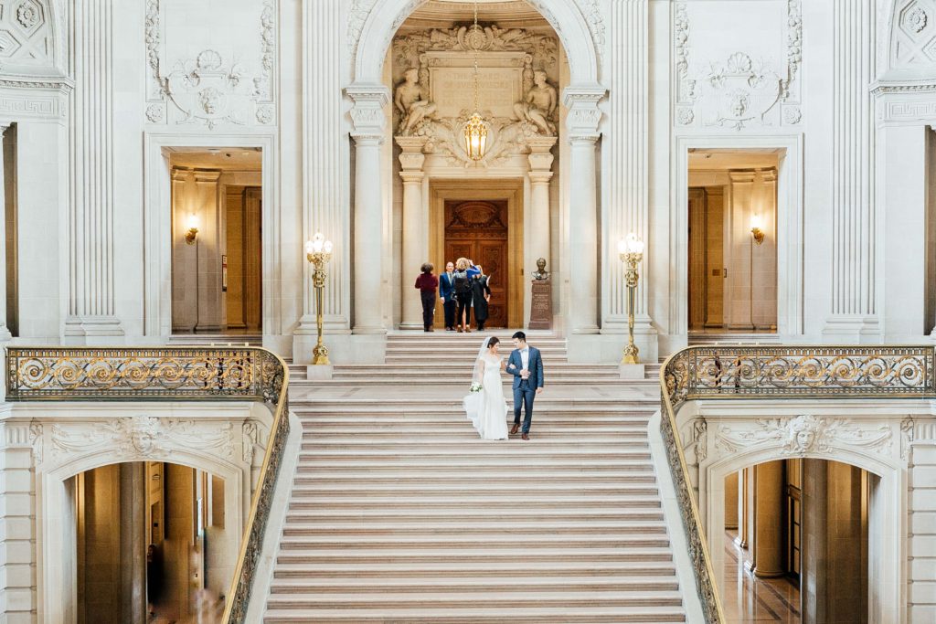 Zoomed out shot of bride and groom walking down main stairs of City Hall