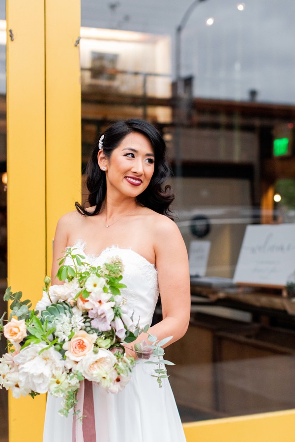 The bride poses for the camera in front of San Francisco wedding venue, stable cafe. She holds her ethical wedding bouquet and smiles.