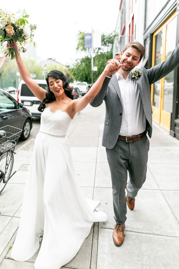 The bride and groom raise their hands up as they walk down a street toward their sustainable wedding in san francisco.