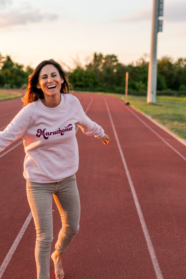 Janel runs on a running track wearing one of her t-shirt designs, a sweatshirt that says "Marathoner"
